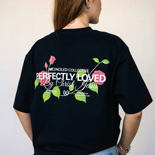 Female model wearing black Christian t-shirt featuring colorful floral “Perfectly Loved By Christ Jesus” back design.