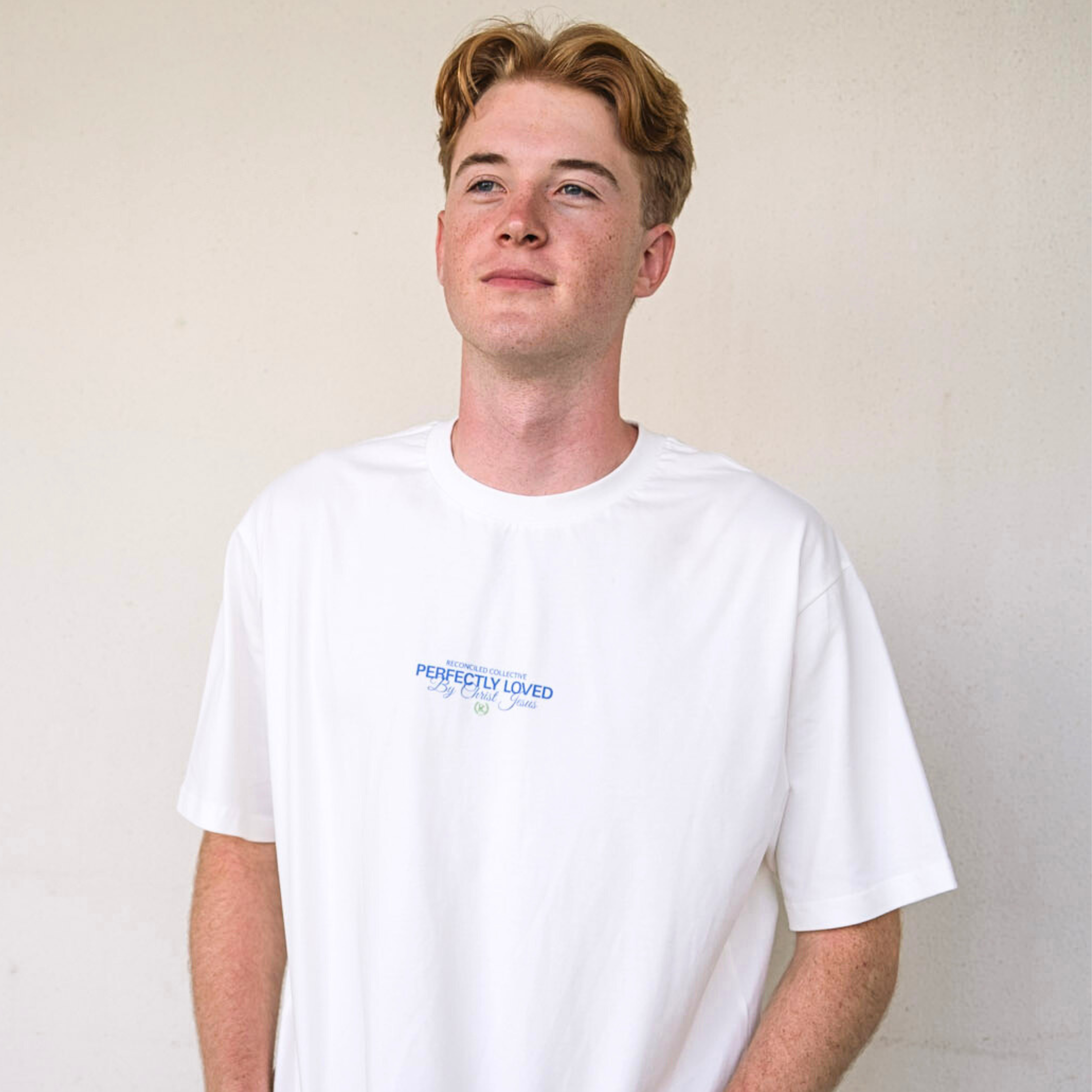 Model wearing white Perfectly Loved Christian streetwear t-shirt by Reconciled Collective with minimalist blue chest print.